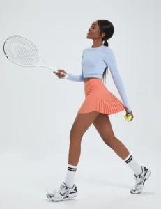 A woman wearing a light coral BALEAF pleated tennis skirt, styled with a comfortable high-waisted fit and built-in shorts, perfect for tennis or pickleball matches.