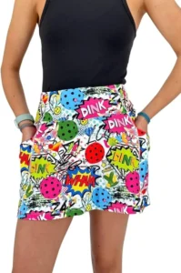 A woman wearing a vibrant comic-style pickleball skirt featuring colorful designs with words like "Dink" and "Wham," equipped with deep side pockets and built-in shorts, perfect for casual and fun pickleball games.